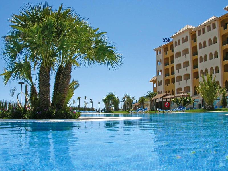Großer blauer Pool mit Palmen und einem mehrstöckigen Hotelgebäude bei klarem Himmel.