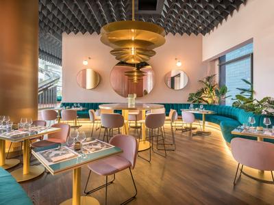 Area ristorante moderna con tavoli rotondi, sedie rosa e divanetti blu sotto lampade sospese dorate.