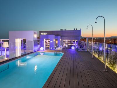 Schwimmbad auf Dachterrasse mit Holzdeck, Glasgeländer und moderner Beleuchtung bei Dämmerung.
