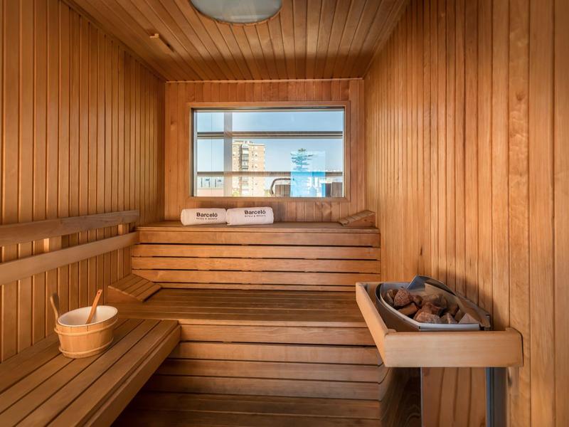 Kleine Sauna mit hellen Holzbohlen, zwei Holzbänken, einem Eimer und Saunaofen mit Steinen.