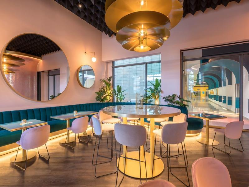 Modernes Hotelrestaurant mit runden Tischen, rosa Stühlen und großen goldenen Deckenlampen.