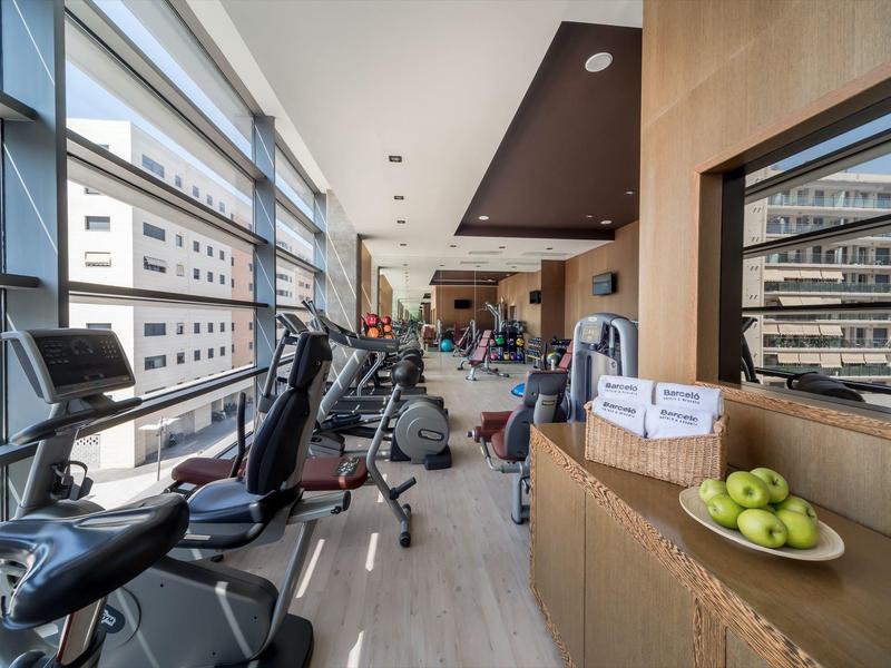 Moderner Fitnessraum mit Cardiogeräten, Fensterfront, hellem Boden und Obstschale auf Sideboard.