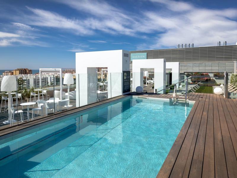 Moderner Außenpool mit klarem Wasser, Holzdeck und weißem Gebäude vor blauem Himmel.