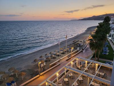 Strand mit Sonnenschirmen und Palmen, ruhiges Meer, Sonnenuntergang, Promenade und Sitzbereiche.