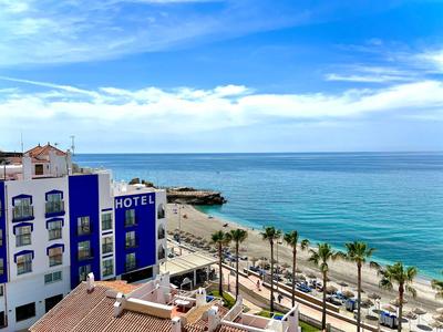 Hotelgebäude mit blauen Akzenten neben einer Palmenallee am Meer unter blauem Himmel.
