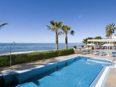 Ein klarer Pool mit blauer Wasserfläche neben Palmen und Meer unter klarem Himmel.