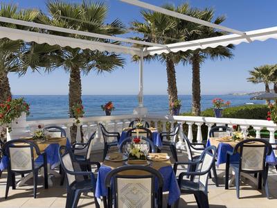 Terrassenrestaurant mit blauen Stühlen, Tischen und Palmen, Blick aufs blaue Meer bei klarem Himmel.