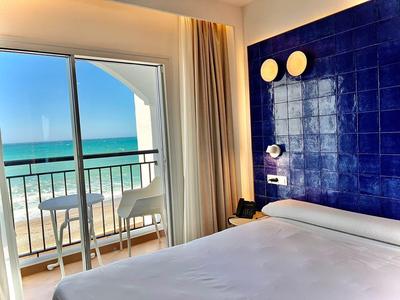 Hotelzimmer mit weißem Bett, blauer Wand und Balkon mit Meerblick bei Sonnenschein.