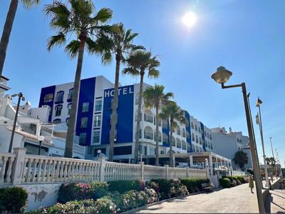 Blauer Himmel über weißem Hotel mit blauen Akzenten, Palmen und Blumen entlang eines Gehwegs.