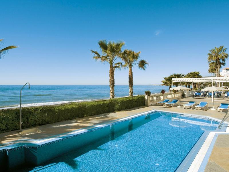 Schwimmbad mit klar blauem Wasser, Palmen, Liegestühlen und Blick auf das ruhige Meer unter blauem Himmel.