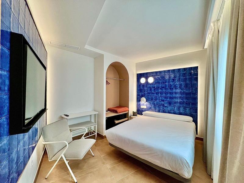 Schlafzimmer mit weißem Bett, blau gekachelter Wand, Fenster mit Gardinen und moderner Einrichtung.