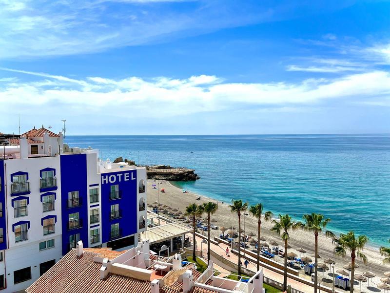 Hotelgebäude mit blauen Akzenten neben einer Palmenallee am Meer unter blauem Himmel.