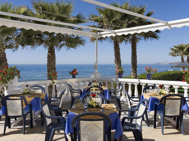 Terrassenrestaurant mit blauen Stühlen, Tischen und Palmen, Blick aufs blaue Meer bei klarem Himmel.