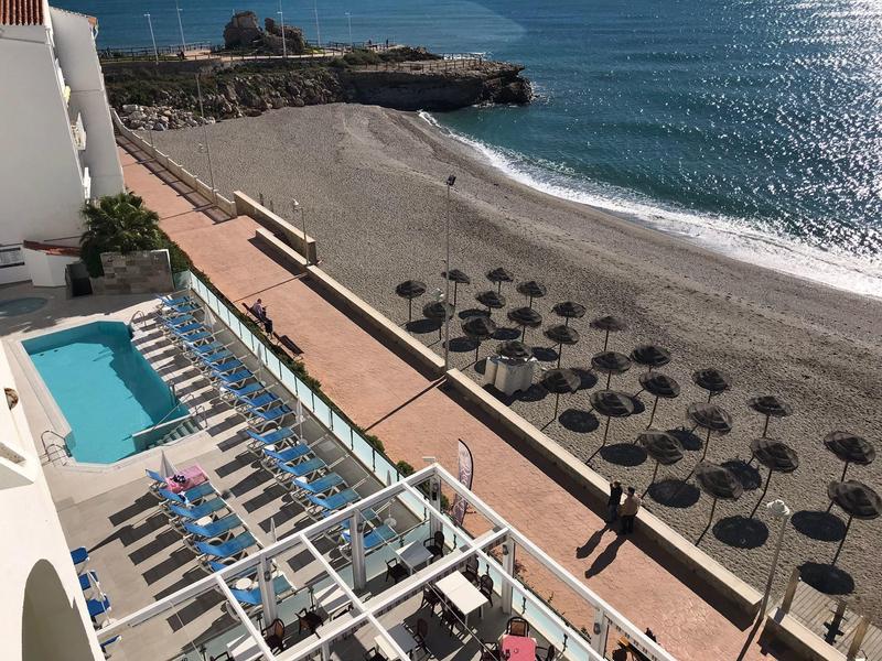 Blick auf einen Strand mit Sonnenschirmen und angrenzendem Hotelpool unter blauem Himmel.
