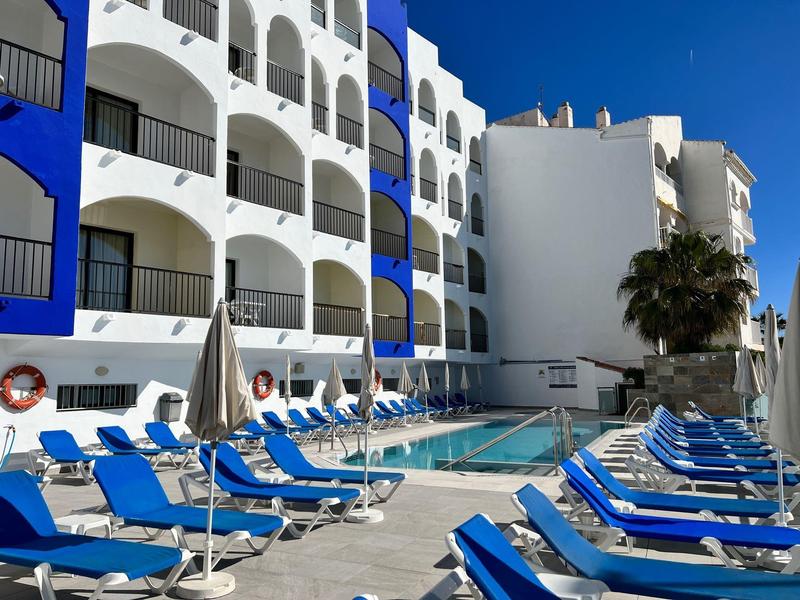 Moderner Hotelpool mit blauen Liegestühlen, weißen Mauern und klar blauem Himmel.
