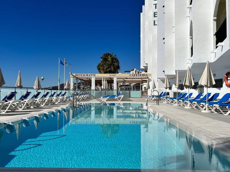 Großer Pool mit klarem Wasser, umgeben von Liegestühlen und Sonnenschirmen vor weißem Hotel.