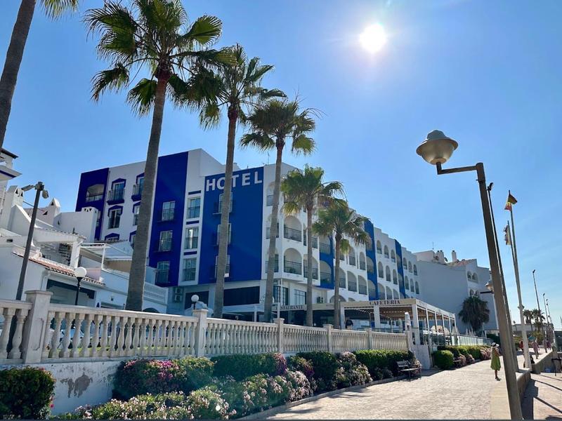 Blauer Himmel über weißem Hotel mit blauen Akzenten, Palmen und Blumen entlang eines Gehwegs.
