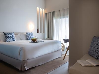 Helles, modernes Schlafzimmer mit großem Bett, blauen Kissen, Vorhängen und schlichtem Dekor.