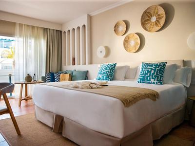 Helles Schlafzimmer mit großem Bett, türkis gemusterten Kissen, Wanddeko aus Körben und Balkonlicht.