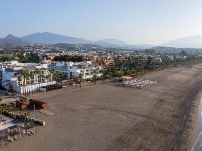 Langer breiter Sandstrand mit verstreuten Sonnenschirmen und Gebäuden am Rand, Berge im Hintergrund.