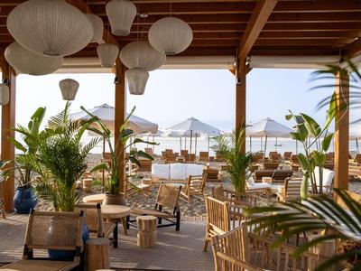 Offenes Strandrestaurant mit Holzdach, runden Lampen, Pflanzen und Blick auf Sand und Meer.