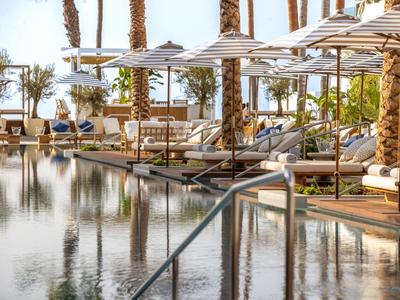 Pool mit ruhigem Wasser, Liegen und Sonnenschirmen, Palmen und Menschen im Hintergrund.