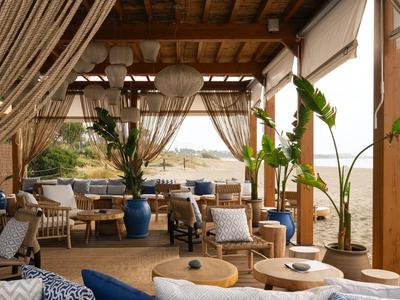 Offene, helle Lounge mit Holzdach, blauen Sitzmöbeln, Pflanzen und Blick auf das Meer.