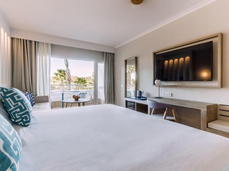 Helles Schlafzimmer mit großem Bett, TV an der Wand, Schreibtisch und Fenster mit Vorhängen.