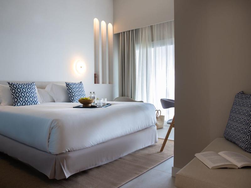 Helles, modernes Schlafzimmer mit großem Bett, blauen Kissen, Vorhängen und schlichtem Dekor.