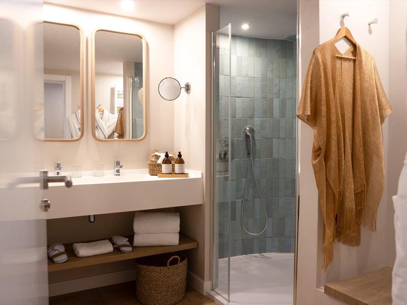 Helles Badezimmer mit Doppelwaschbecken, großem Spiegel, Glasdusche und beige Bademantel am Haken.