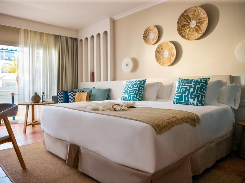 Helles Schlafzimmer mit großem Bett, türkis gemusterten Kissen, Wanddeko aus Körben und Balkonlicht.