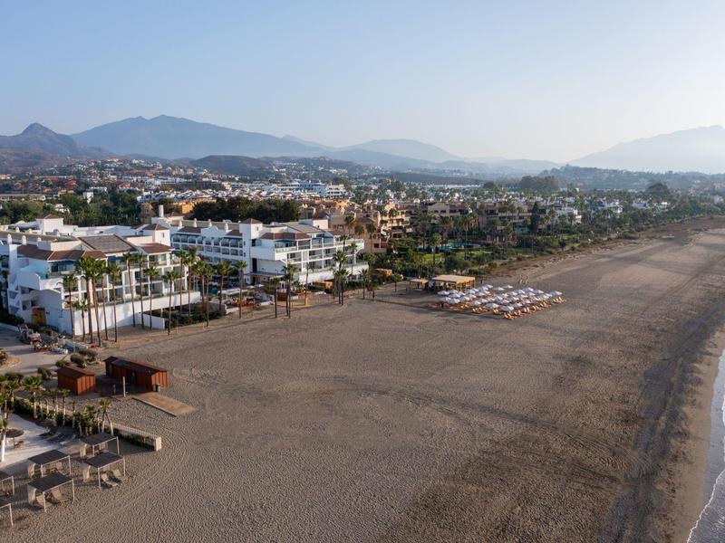 Langer breiter Sandstrand mit verstreuten Sonnenschirmen und Gebäuden am Rand, Berge im Hintergrund.