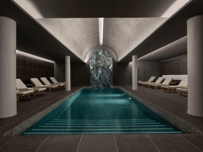 Innenpool in modernem Raum mit hellen Säulen, Liegestühlen und Wasserfall-Farbspiel am Ende.