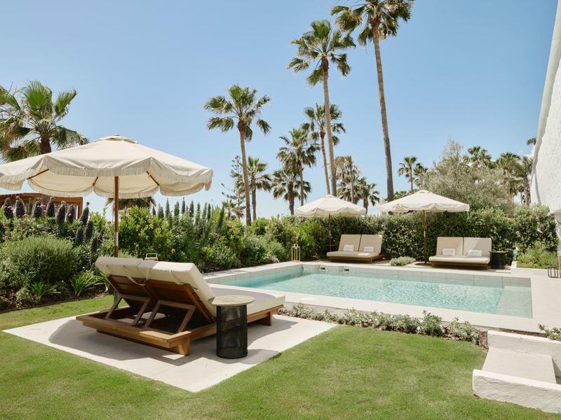 Schwimmbecken mit Liegen und Sonnenschirmen, umgeben von Palmen und grünem Rasen.