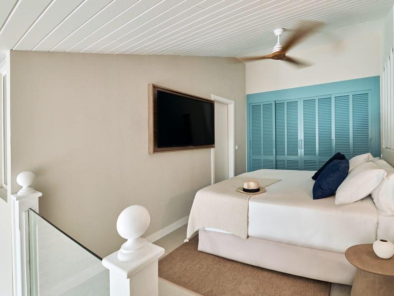 Helles Schlafzimmer mit Doppelbett, blauen Kissen, Ventilator und Fernseher an weißer Wand.