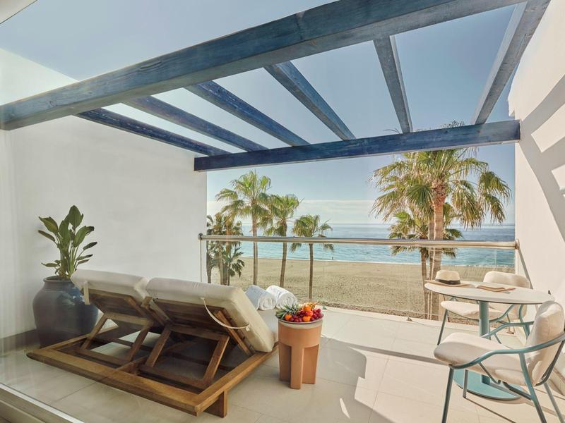 Balkon mit blauen Holzbalken, Liegestühlen und Meerblick, Palmen im Hintergrund.