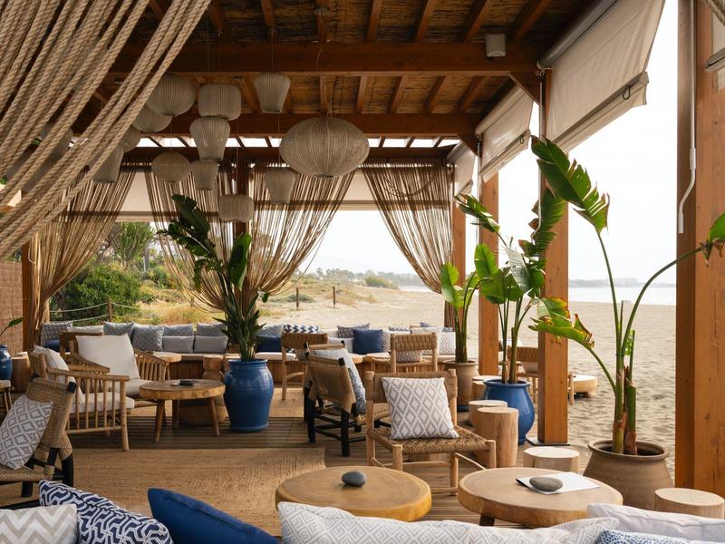 Offene, helle Lounge mit Holzdach, blauen Sitzmöbeln, Pflanzen und Blick auf das Meer.