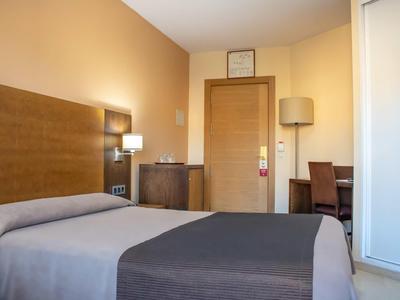 Schlichtes Hotelzimmer mit Einzelbett, hellem Holz, grauer Bettdecke und Schreibtisch im Hintergrund.