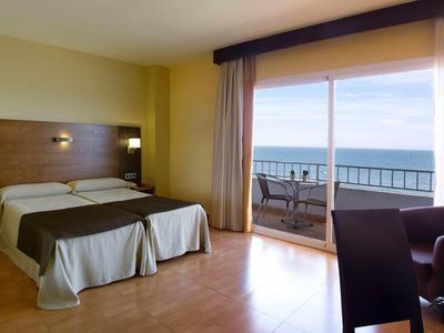 Helles Hotelzimmer mit Doppelbett, Holzboden, Balkon und Meerblick bei Tageslicht.