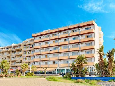 Sechs Stockwerke hohes Wohngebäude mit Balkonen, Palmen und blauem Himmel im sonnigen Umfeld.