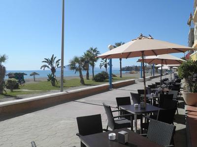 Straßenansicht mit Café-Tischen und Stühlen unter Sonnenschirmen, Palmen und Meer im Hintergrund.