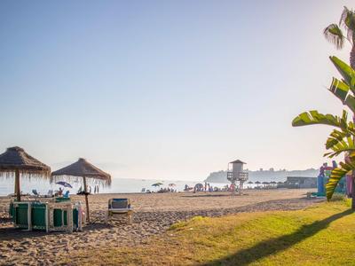 Strand mit Sand, Strohhütten, Sonnenliegen, Palmen und weiter Meeresblick bei klarem Himmel.