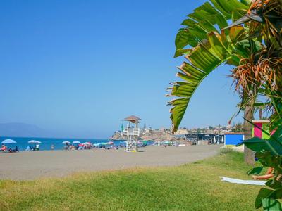 Strand mit blauem Himmel, Sand, Sonnenschirmen und Palmenblättern rechts im Vordergrund.