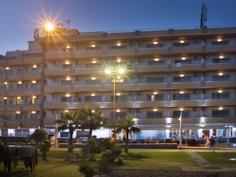 Mehrstöckiges Hotel mit Balkonreihen, beleuchtet bei Dämmerung, davor grasbewachsenes Gelände mit Palmen.