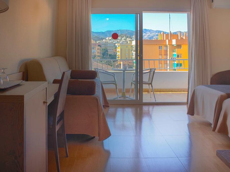 Helles Hotelzimmer mit zwei Betten, Tisch, Stühlen und Balkon mit Stadt- und Bergblick.