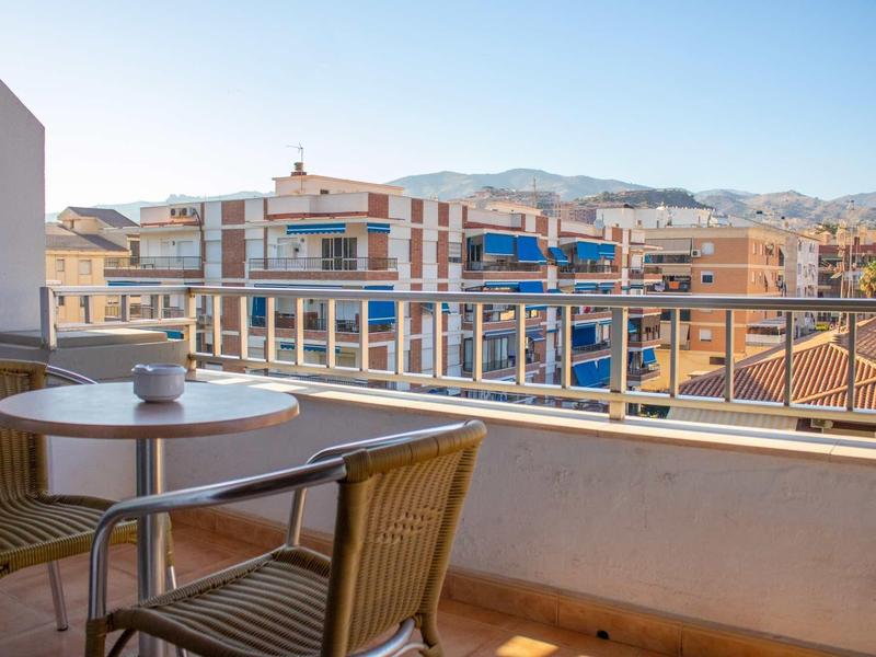 Balkon mit rundem Tisch, zwei Stühlen und Blick auf Wohnhäuser und Berge im Hintergrund.