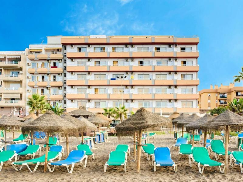 Ein Hotel mit mehreren Stockwerken, davor viele blaue Liegestühle und Strohdächer am Strand.
