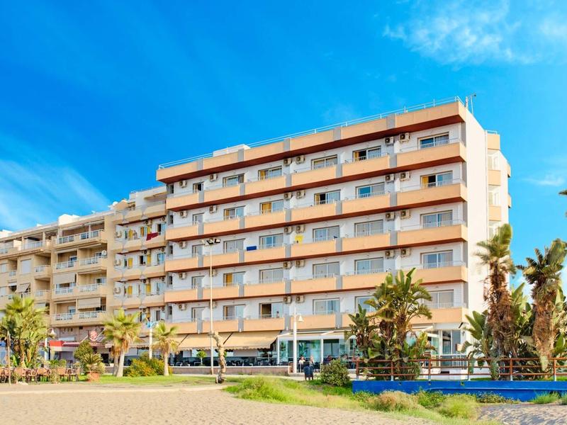 Sechs Stockwerke hohes Wohngebäude mit Balkonen, Palmen und blauem Himmel im sonnigen Umfeld.