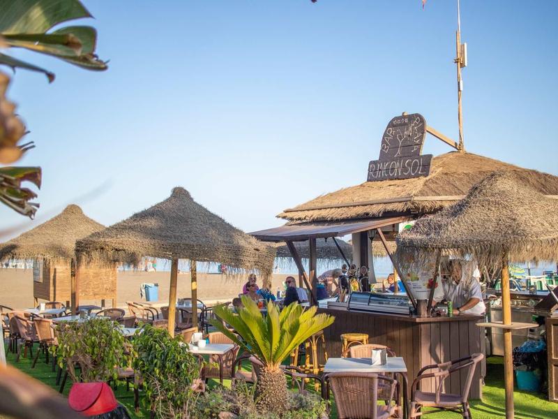 Strandbar mit Strohdächern und Sonnenschirmen, blauer Himmel und grünen Pflanzen im Vordergrund.