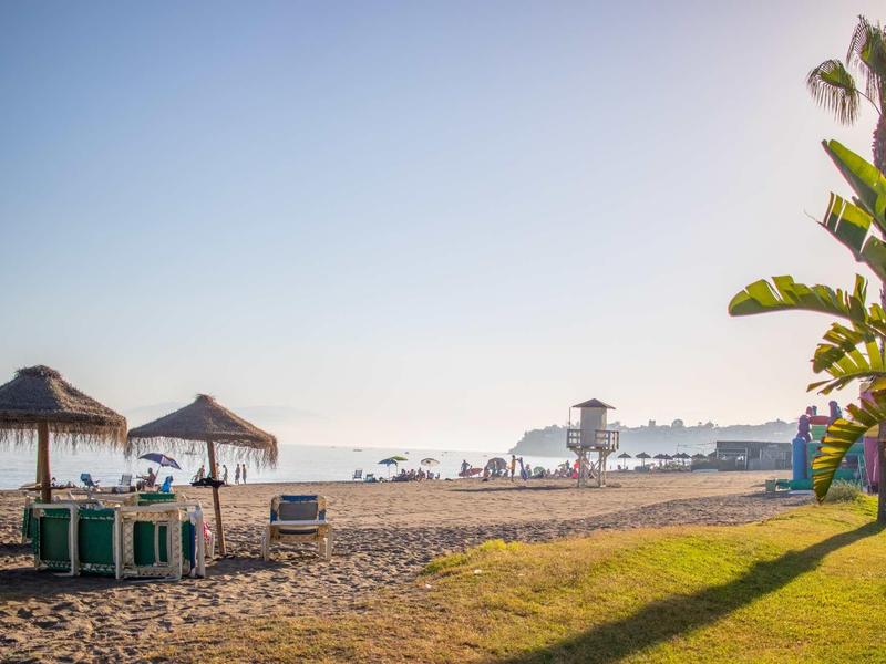 Strand mit Sand, Strohhütten, Sonnenliegen, Palmen und weiter Meeresblick bei klarem Himmel.